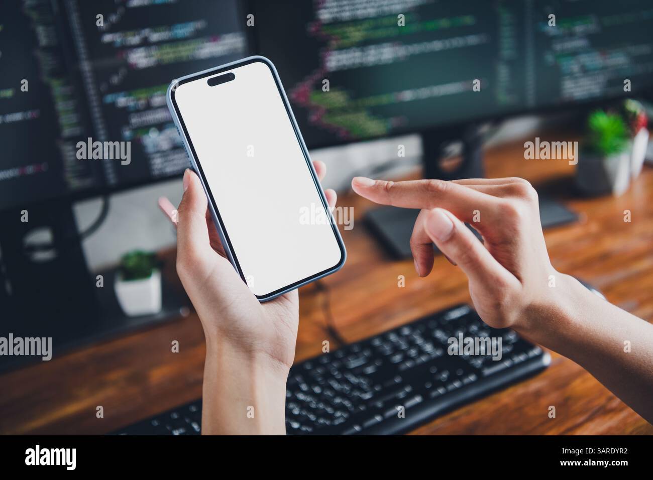 Giovane sviluppatrice di software donna che utilizza lo smartphone per la codifica in uno spazio di lavoro moderno con schermi di computer e strumenti di programmazione Foto Stock