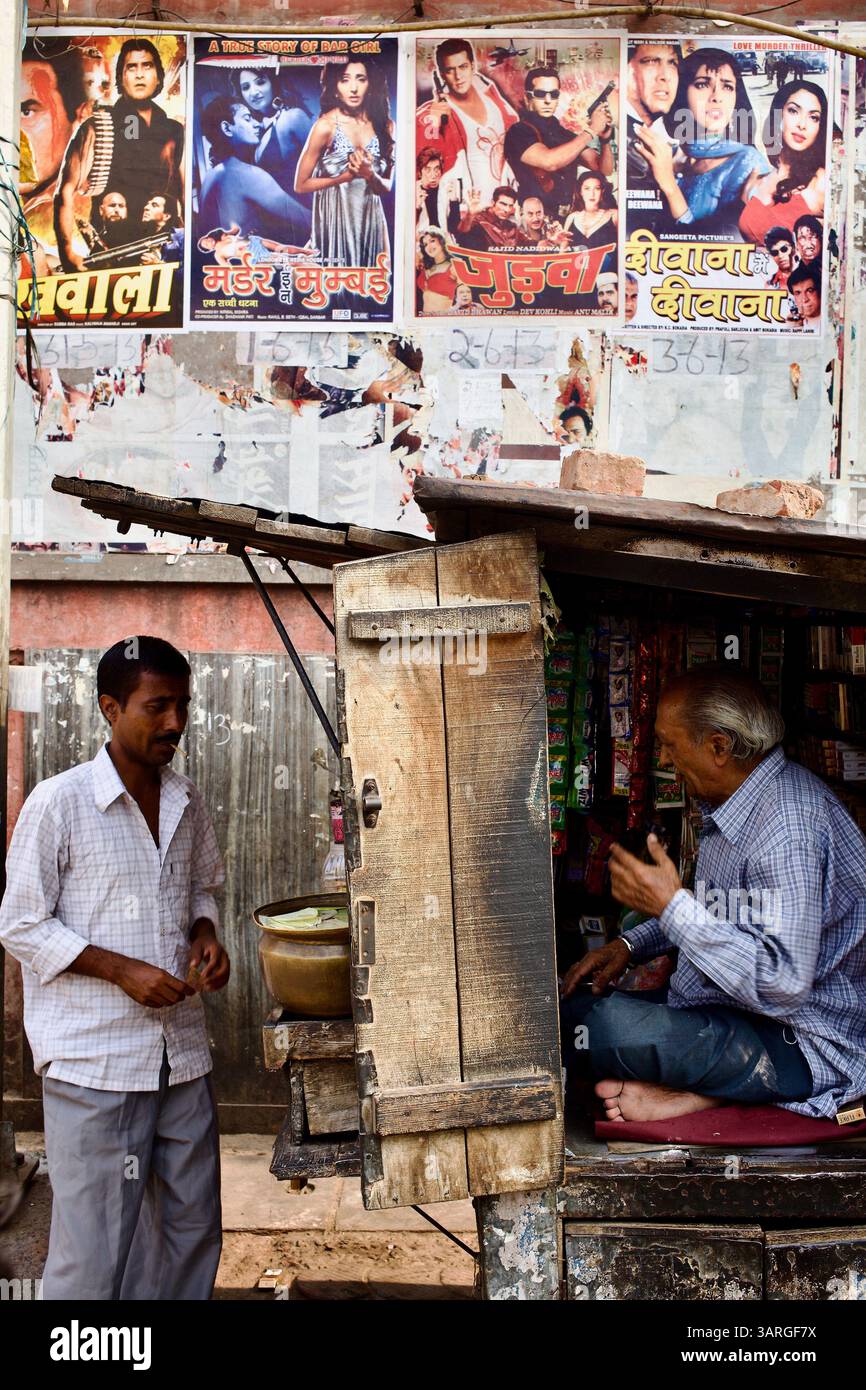 Un venditore di strada chatta con un negoziante all'interno di un chiosco di legno sotto i vecchi poster dei film di Bollywood su un muro di sbucciatura a Delhi, in India. Foto Stock