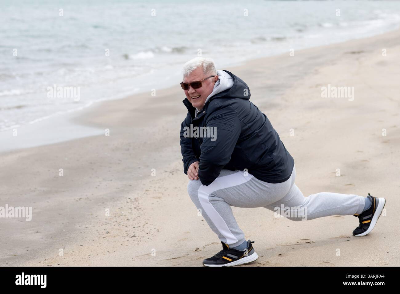 Uomo anziano che si allunga su una spiaggia fredda, abbracciando il fitness all'aperto con energia e gioia Foto Stock