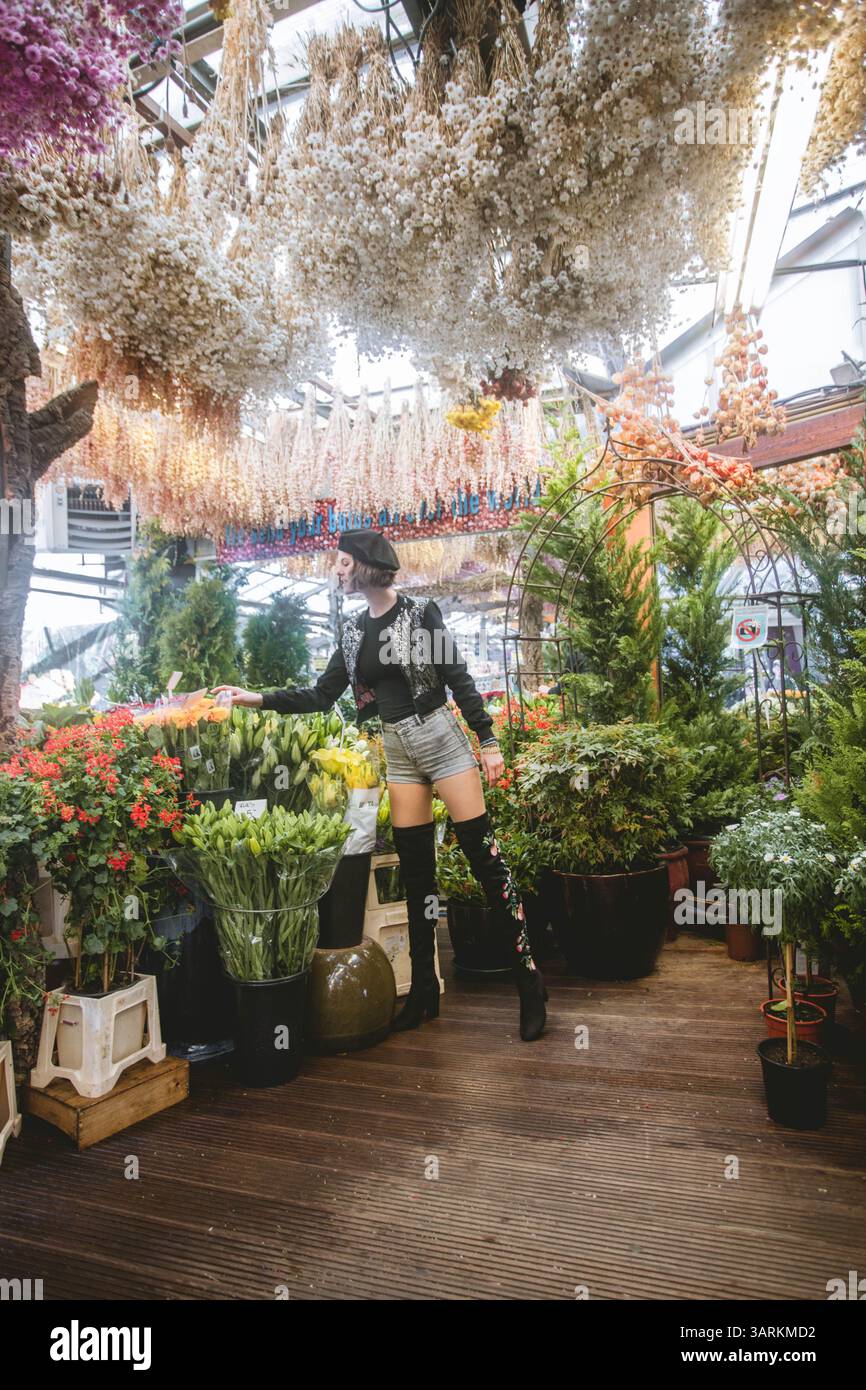 Bella giovane donna che indossa pantaloni corti, bomber corto e stivali neri alti. La modella di moda sta godendo la bellezza e l'odore dei fiori colorati Foto Stock