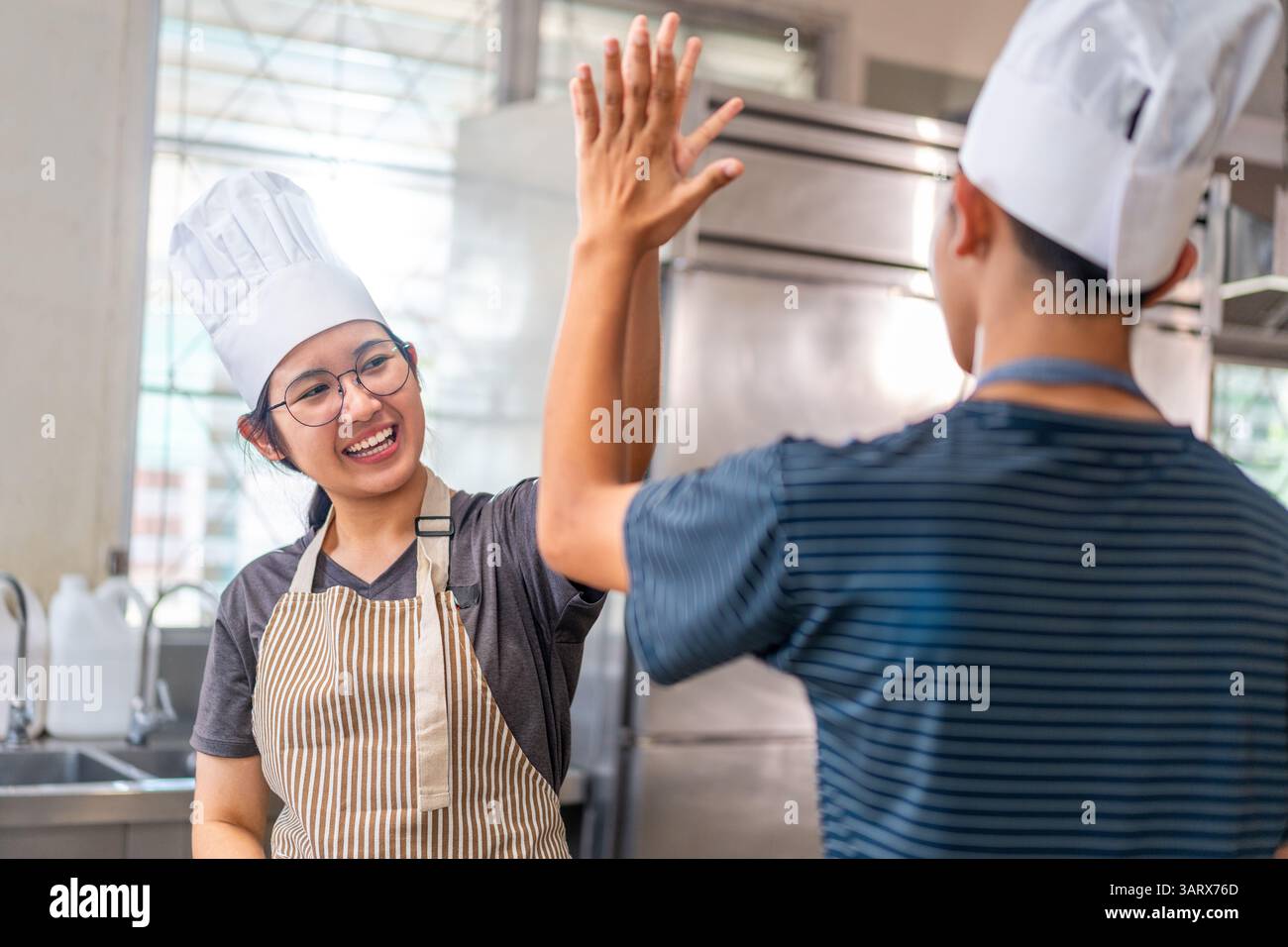 Due giovani chef festeggiano con un alto cinque in una cucina commerciale, indossando cappelli e grembiuli da chef, mostrando lavoro di squadra e gioia nella cucina. Foto Stock