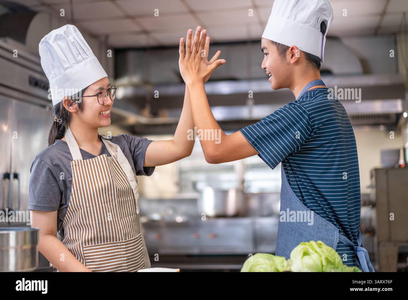 Due giovani chef festeggiano con un alto cinque in una cucina commerciale, indossando cappelli e grembiuli da chef, mostrando lavoro di squadra e gioia nella cucina. Foto Stock