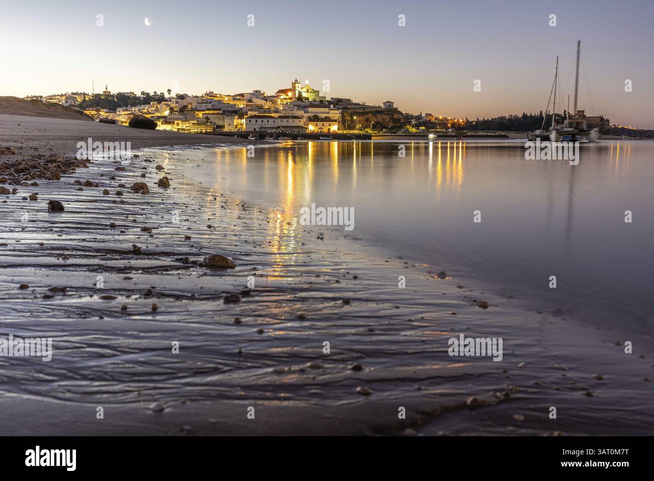 Ripresa notturna di una spiaggia sabbiosa e del porto con la bassa marea. Immagine panoramica di un centro storico illuminato all'alba a Ferragudo, Portimao, Algarve, Foto Stock