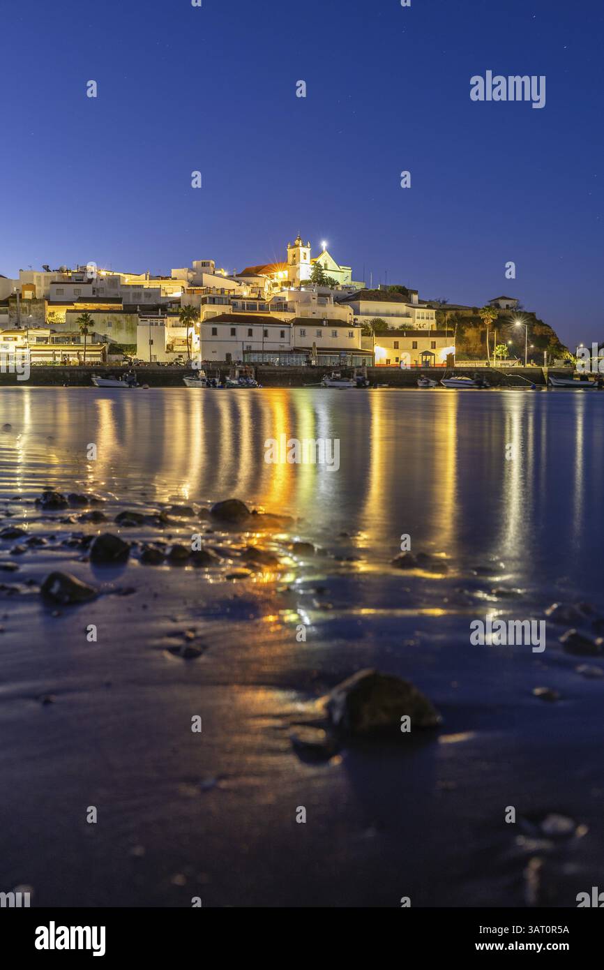 Ripresa notturna di una spiaggia sabbiosa e del porto con la bassa marea. Immagine panoramica di un centro storico illuminato all'alba a Ferragudo, Portimao, Algarve, Foto Stock