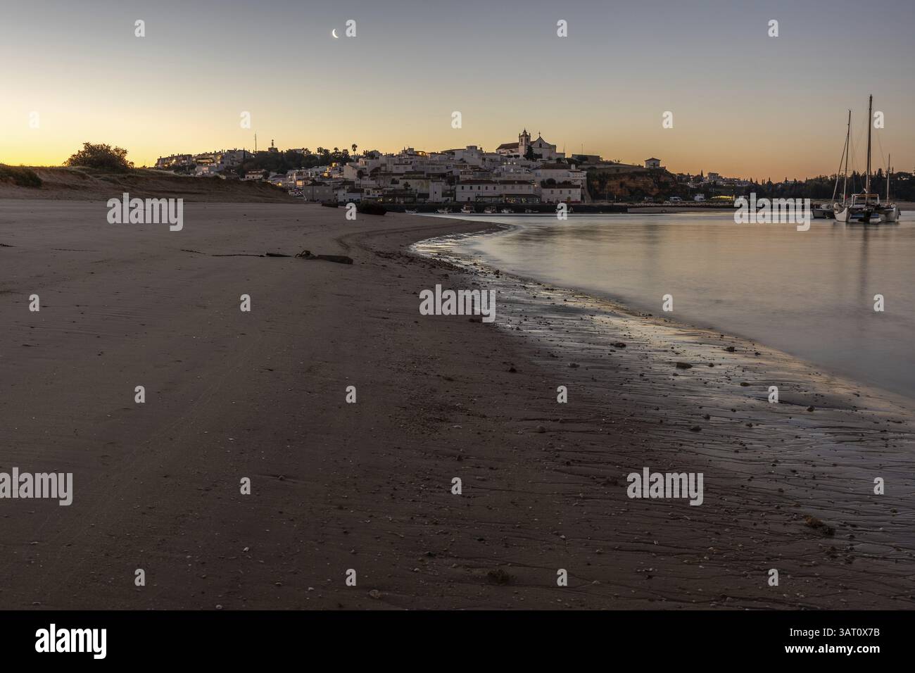 Ripresa notturna di una spiaggia sabbiosa e del porto con la bassa marea. Immagine panoramica di un centro storico illuminato all'alba a Ferragudo, Portimao, Algarve, Foto Stock