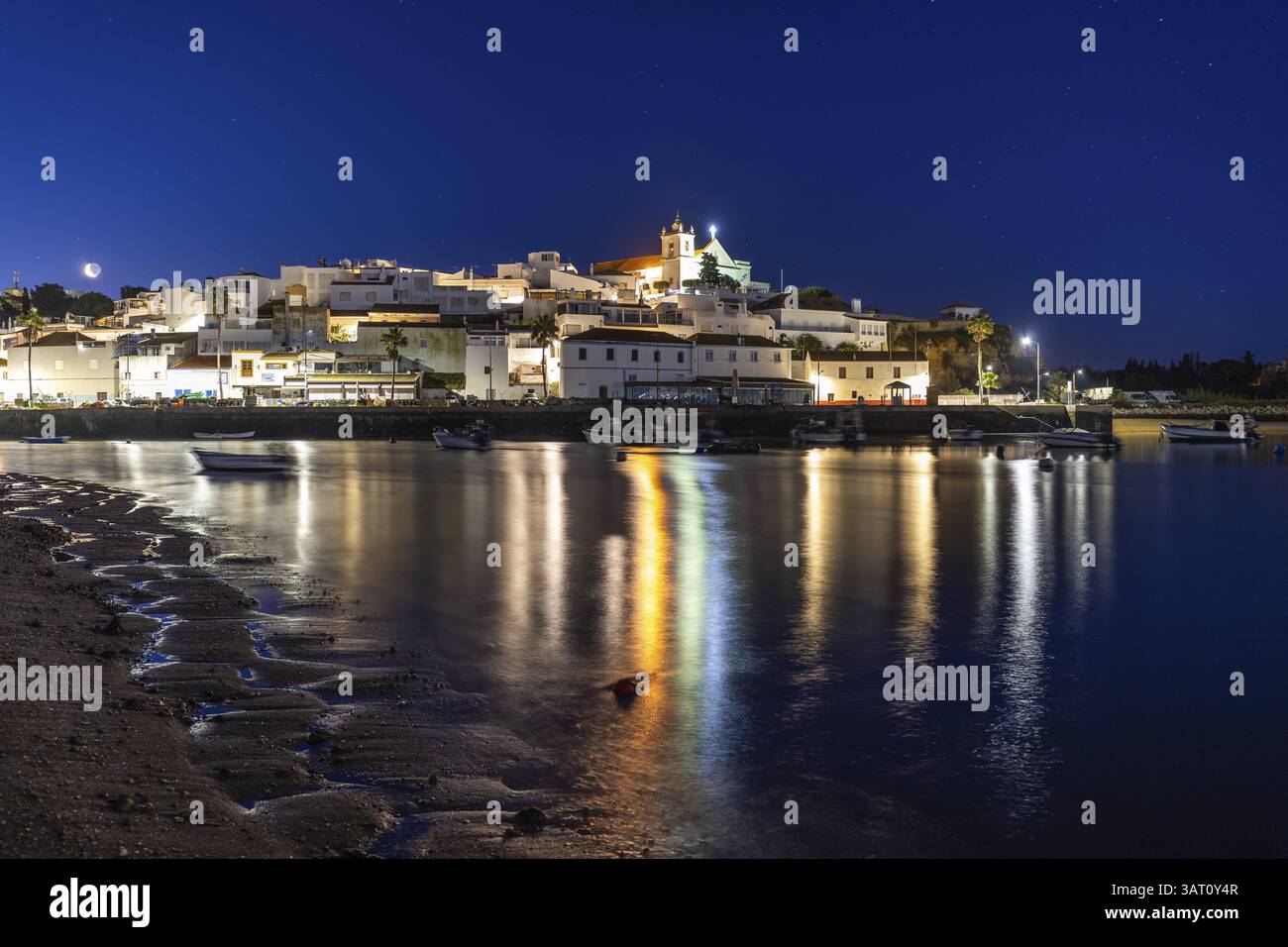 Ripresa notturna di una spiaggia sabbiosa e del porto con la bassa marea. Immagine panoramica di un centro storico illuminato all'alba a Ferragudo, Portimao, Algarve, Foto Stock