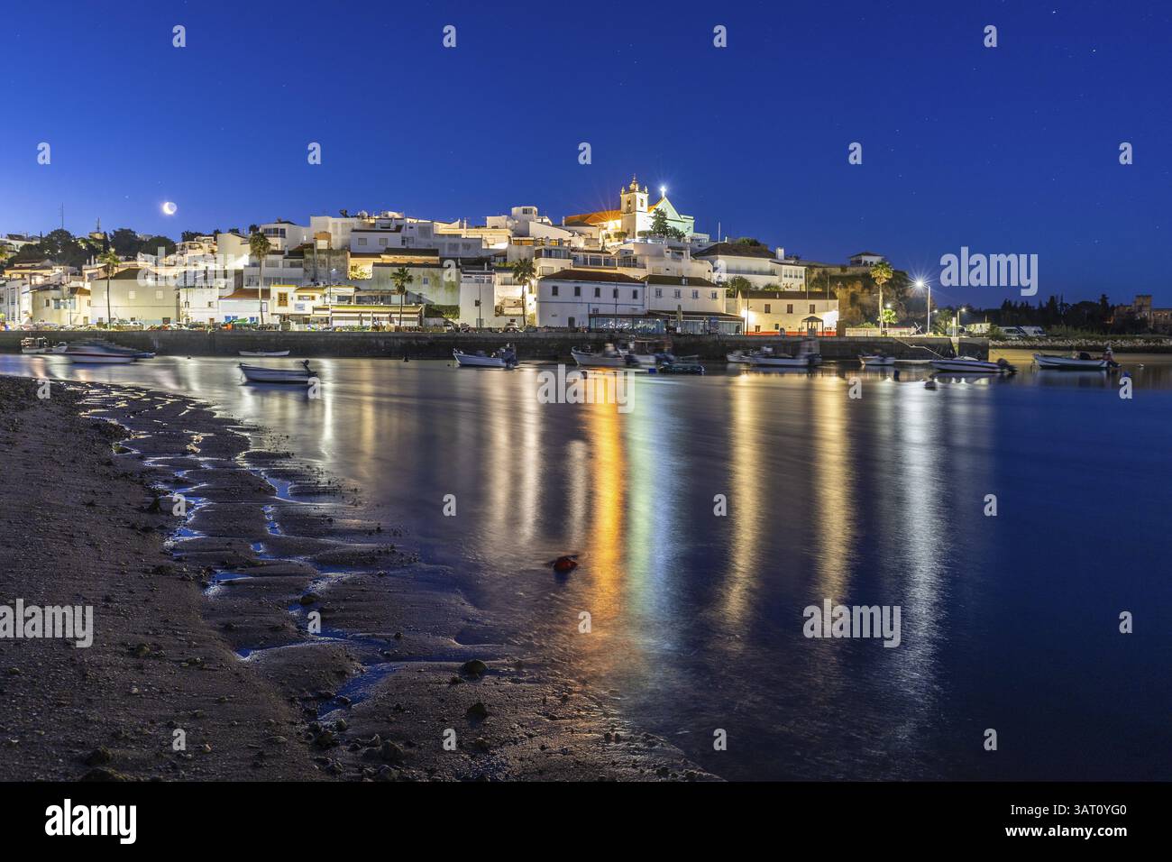 Ripresa notturna di una spiaggia sabbiosa e del porto con la bassa marea. Immagine panoramica di un centro storico illuminato all'alba a Ferragudo, Portimao, Algarve, Foto Stock
