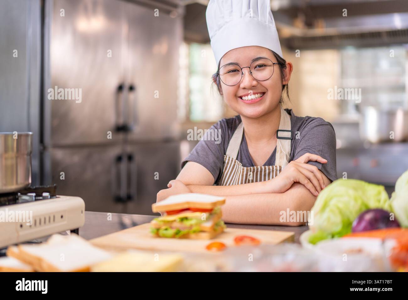 Una giovane e allegra chef donna che indossa un cappello bianco e un grembiule a righe sorride mentre riposa le braccia sul tavolo in una cucina commerciale. Di fronte a lui Foto Stock