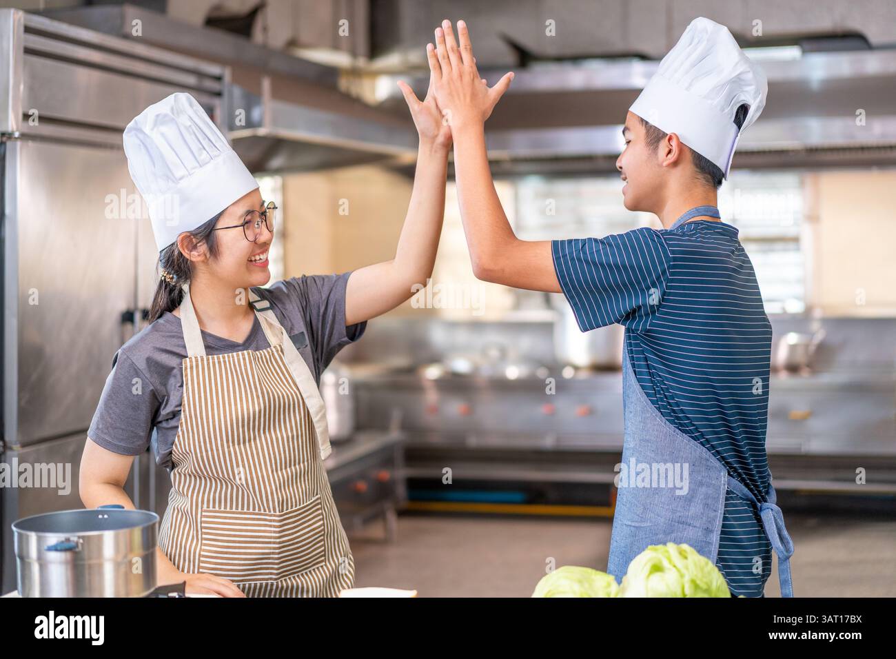 Due giovani chef festeggiano con un alto cinque in una cucina commerciale, indossando cappelli e grembiuli da chef, mostrando lavoro di squadra e gioia nella cucina. Foto Stock