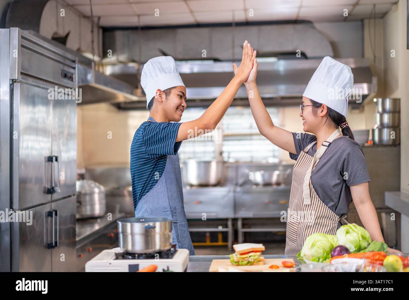 Due giovani chef festeggiano con un alto cinque in una cucina commerciale, indossando cappelli e grembiuli da chef, mostrando lavoro di squadra e gioia nella cucina. Foto Stock