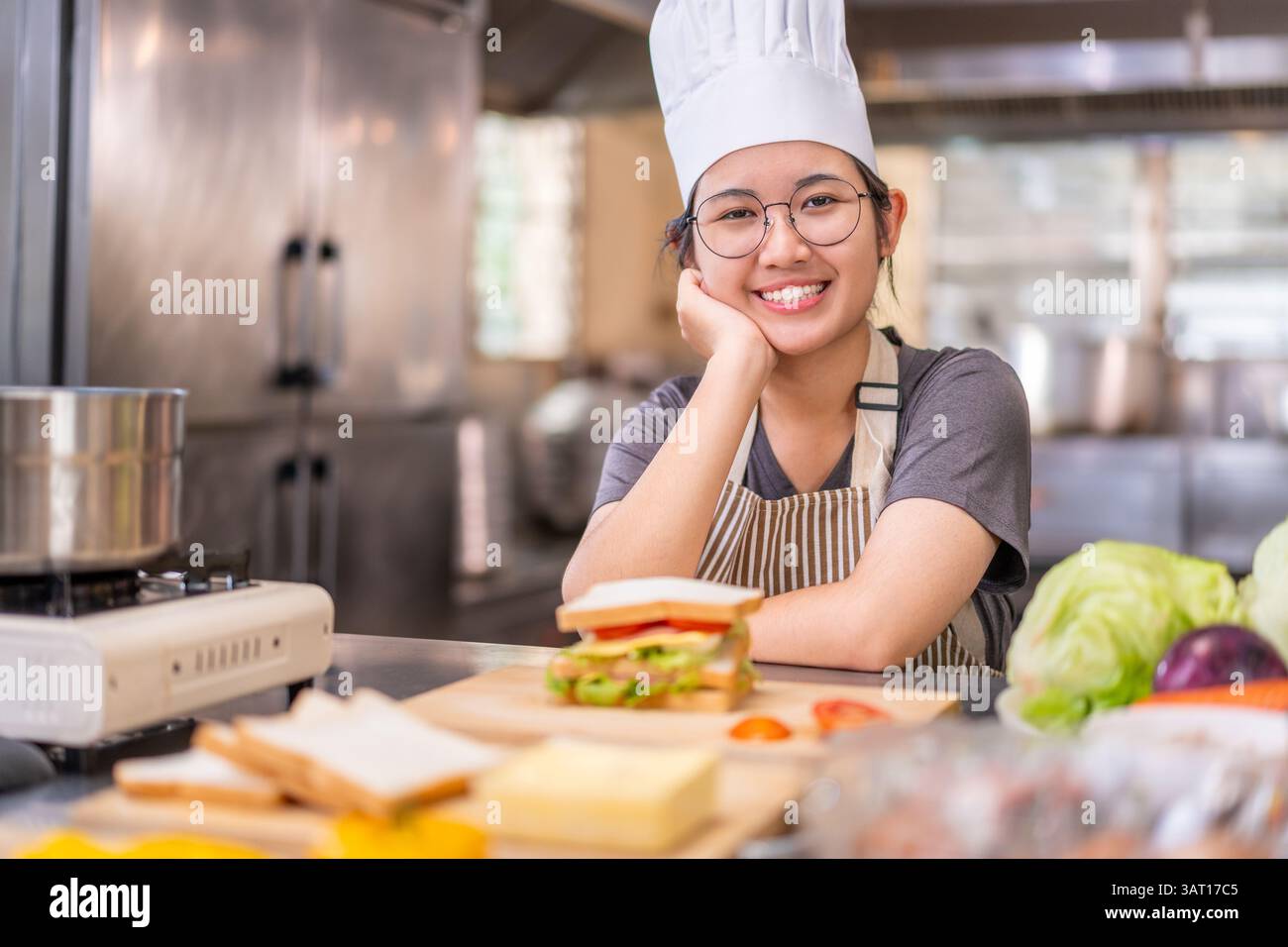 Una giovane e allegra chef donna che indossa un cappello bianco e un grembiule a righe sorride mentre riposa la testa sulla mano in una cucina commerciale. Davanti a lei Foto Stock