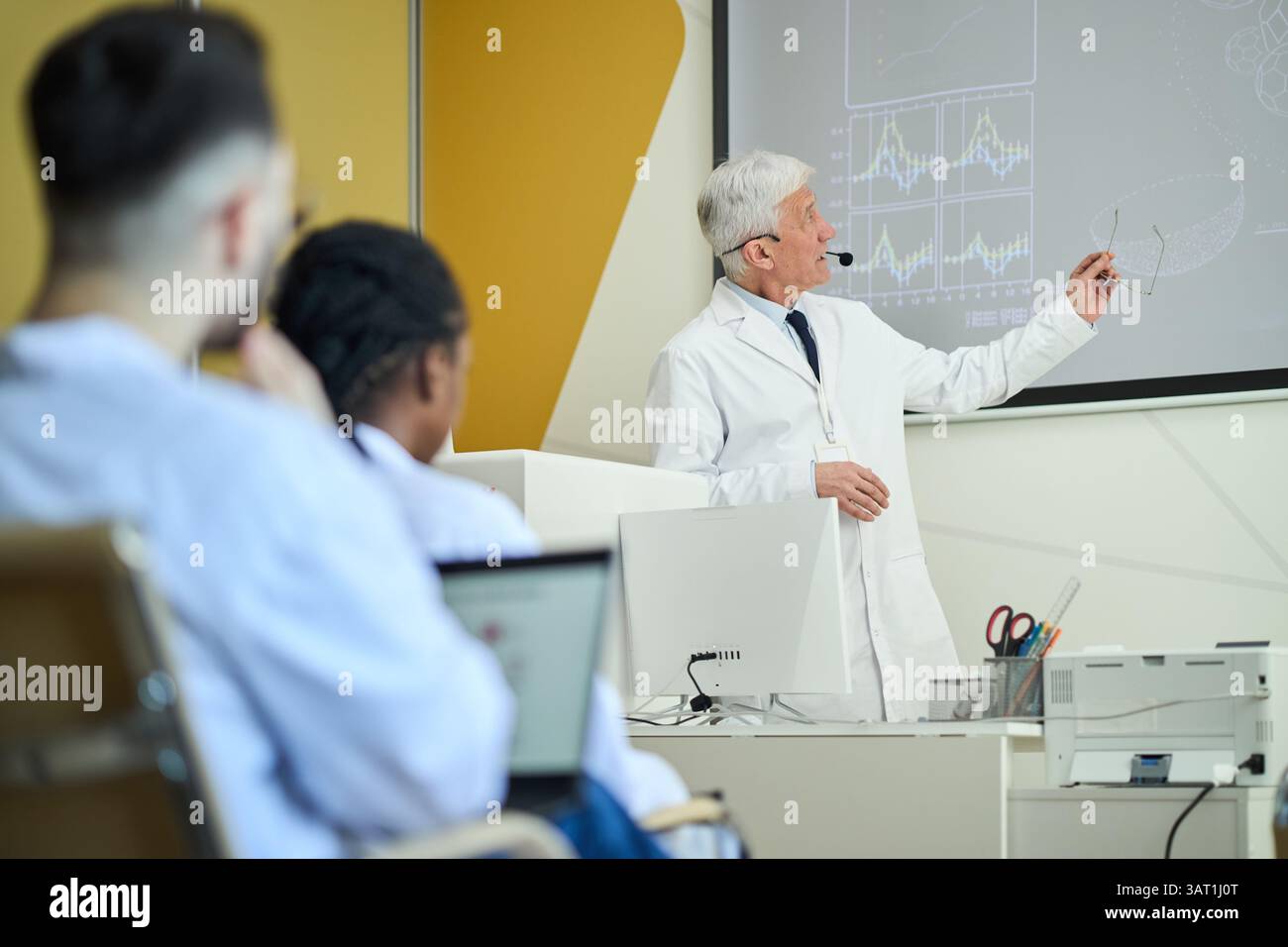 Scienziato senior che tiene lezioni in classe moderna utilizzando schermo e tecnologia, ascoltando e prendendo appunti con attenzione da un pubblico eterogeneo Foto Stock