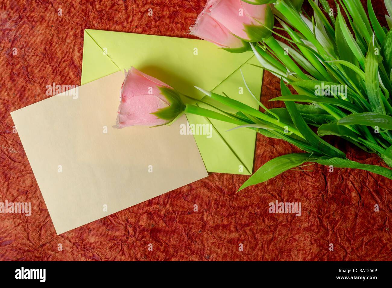Una rosa rosa in busta verde, posizionata su un tessuto testurizzato con un biglietto bianco, è perfetta per i temi di saluto o di comunicazione. Foto Stock