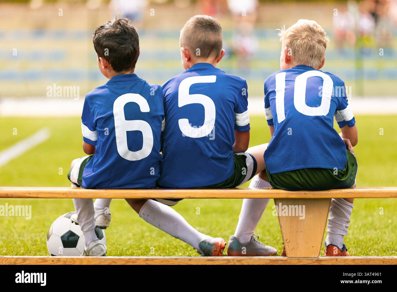 I ragazzi con maglie da calcio blu con numeri bianchi siedono su una panchina di legno sostitutiva, a guardare la partita, uniti come una squadra Foto Stock