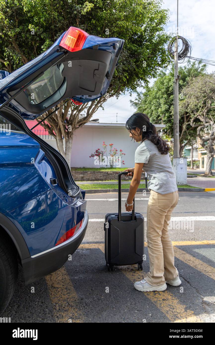 Un adolescente si sta preparando a partire in un'auto blu con una valigia nel bagagliaio. Indossa una camicia grigia e pantaloni abbronzati Foto Stock Un adolescente si sta preparando a partire in un'auto blu con una valigia nel bagagliaio. Indossa una camicia grigia e pantaloni abbronzati Foto Stock