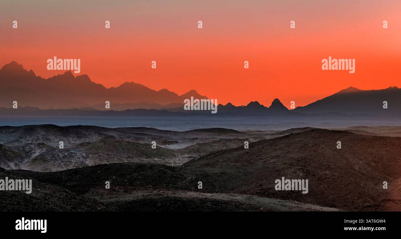 Un paesaggio mozzafiato al tramonto, con montagne a strati sullo sfondo e un vivace cielo arancione. Il primo piano presenta colline ondulate Foto Stock