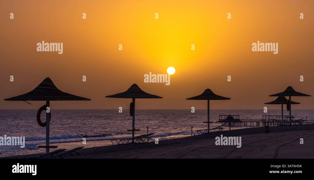 Una tranquilla spiaggia al tramonto, con ombrelloni sagomati contro un vivace cielo arancione. Il sole tramonta sull'oceano calmo, creando un laghetto Foto Stock