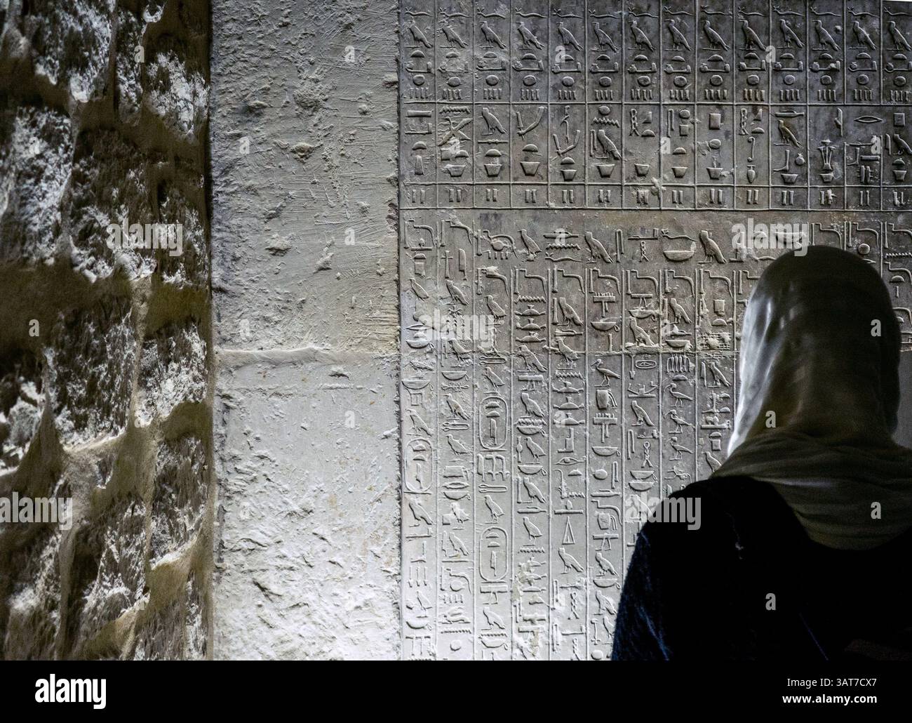 Una persona con un velo si erge di fronte agli antichi geroglifici egiziani scolpiti in un muro di pietra. La consistenza del muro contrasta con lo smoot Foto Stock