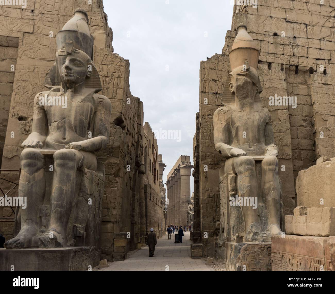 Due grandi statue egiziane che fiancheggiano un sentiero in pietra, con geroglifici sulle pareti e un cielo nuvoloso sopra. I visitatori possono essere visti camminare in t Foto Stock