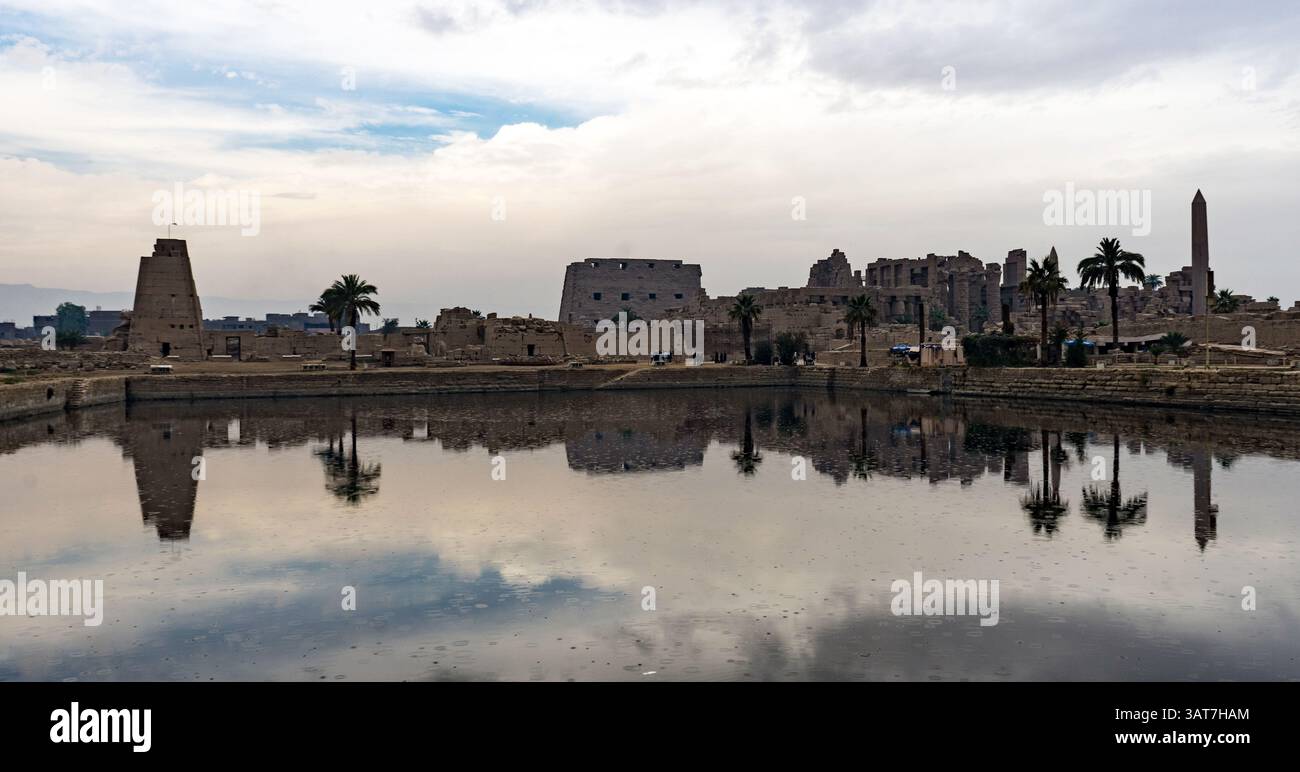 Una vista panoramica delle antiche rovine da un corpo d'acqua calmo, caratterizzato da palme e strutture storiche che si riflettono nell'acqua sotto un cielo nuvoloso. Foto Stock