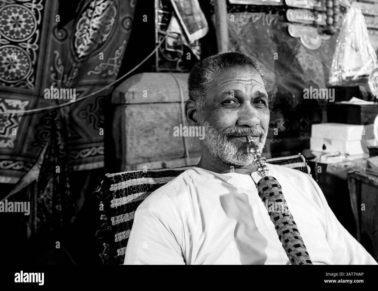 Un uomo di mezza età con la barba seduto in un mercato, che tiene un narghilè tradizionale. Lo sfondo presenta tessuti colorati e vari articoli di mercato, Foto Stock
