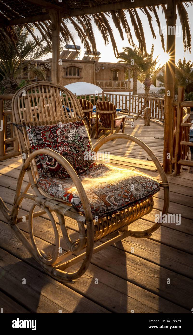 Una rustica sedia a dondolo con un cuscino colorato siede su un ponte di legno, inondata dalla calda luce del sole. Palme e un'atmosfera intima creano un ambiente tranquillo Foto Stock