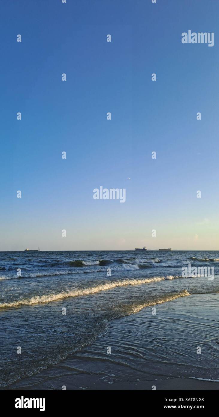 Serena vista costiera delle dolci onde che si infrangono sulla riva sotto un vasto cielo blu nel tardo pomeriggio. Le navi da carico sono visibili all'orizzonte. - Immagine stock catturata con smartphone