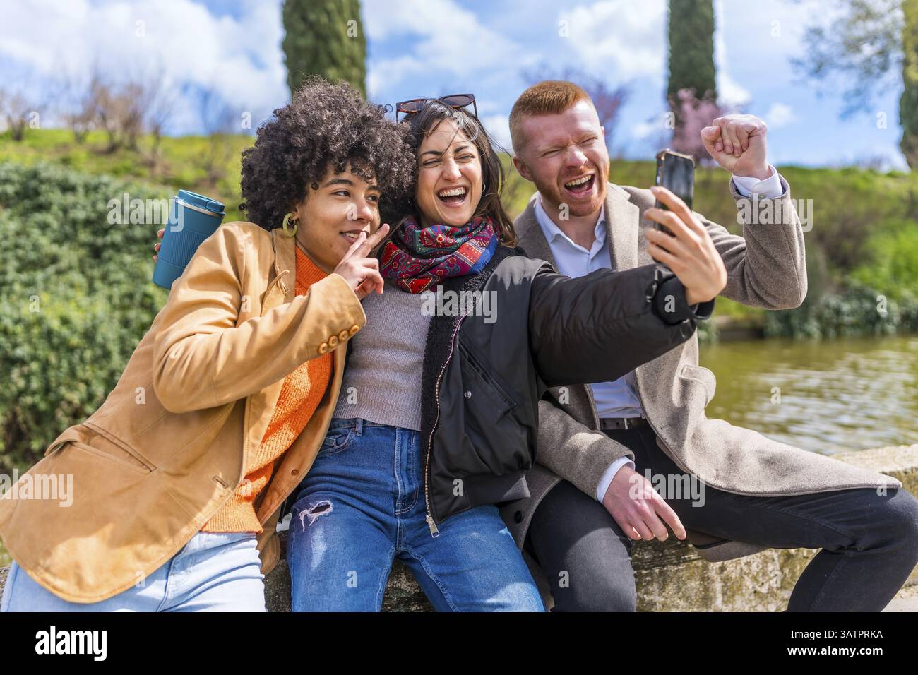 Tre allegri amici multietnici stanno facendo un selfie mentre sono seduti su un muro in un parco vicino a un lago Foto Stock