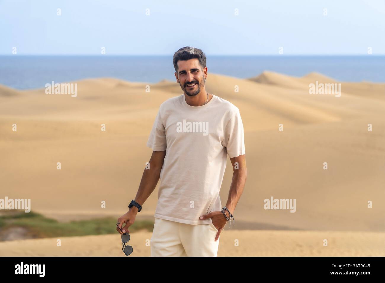 Giovane uomo che si gode una vacanza estiva in mezzo allo splendido paesaggio desertico delle dune di maspalomas, crogiolandosi al sole caldo e al vibrante paesaggio Foto Stock