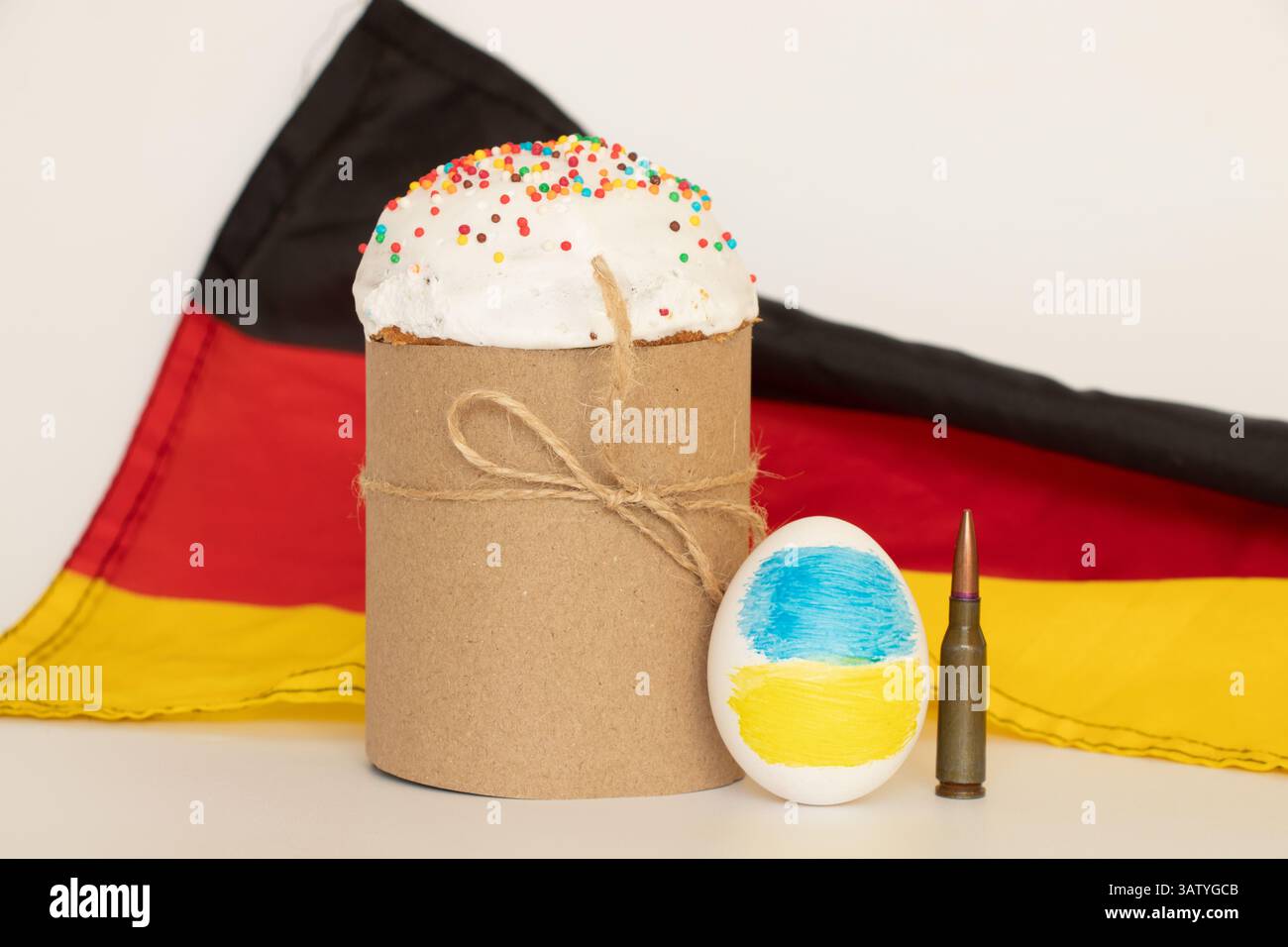 Torta di Pasqua, uovo con bandiera Ucraina e proiettile sullo sfondo della bandiera tedesca sul bianco, aiuti all'Ucraina dalla Germania Foto Stock