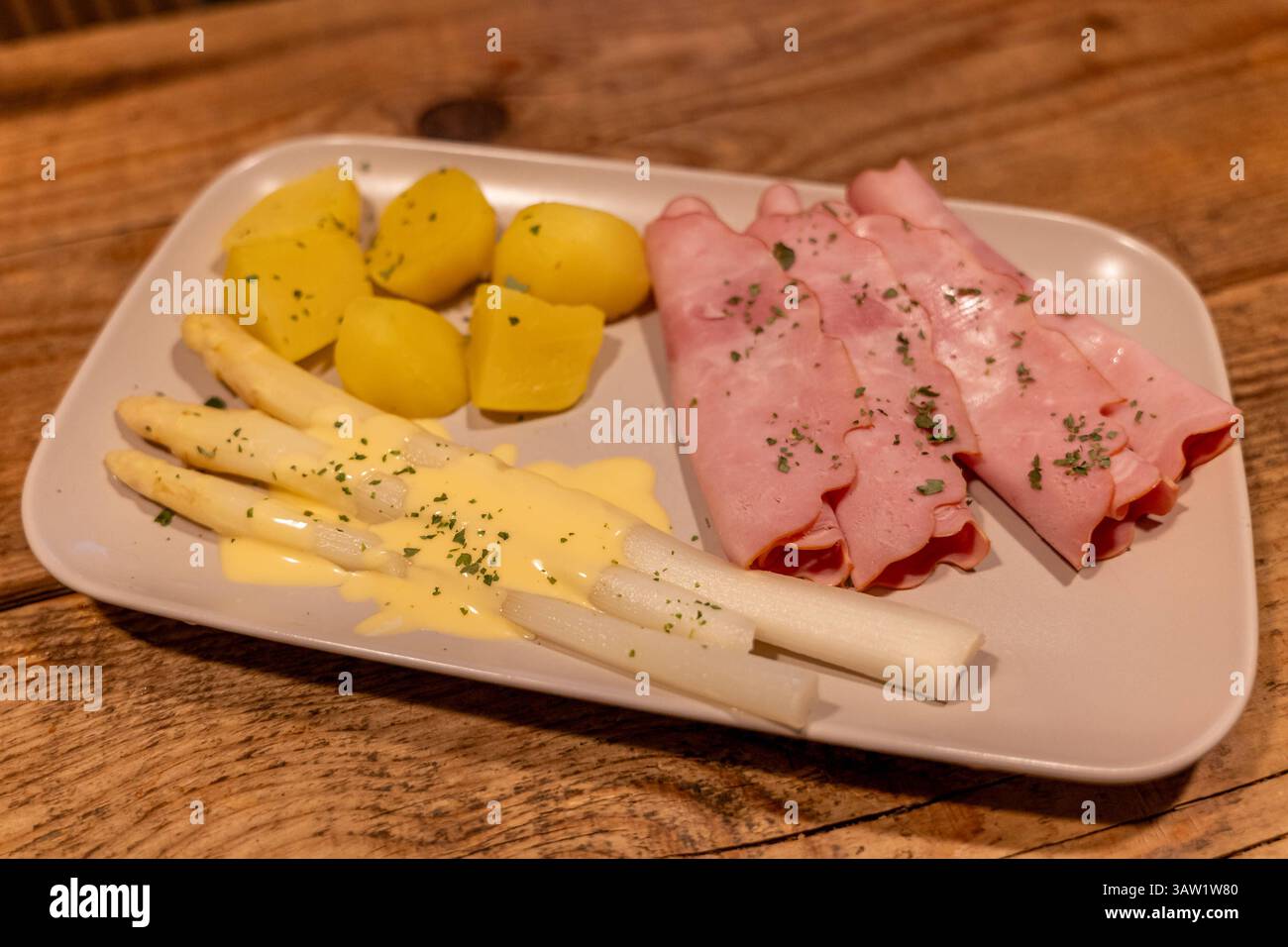 Klassisches Spargelgericht mit Schinken, Kartoffeln und Sauce Hollandaise 17.04.25, Selters: Symbolfoto, Illustrationsbild, Symbolbild, Illustrationsfoto, Alltagsszene Ein traditionelles deutsches Frühlingsgericht mit weißem Spargel, serviert mit Sauce Hollandaise, gekochten Kartoffeln und geräuchertem Kochschinken. DAS Gericht ist angerichtet auf einem hellen rechteckigen Teller auf einem rustikalen Holztisch. Der weiße Spargel ist leicht mit Petersilie garniert, die Kartoffeln sind geschält und in Stücke geschnitten. Eine typische Mahlzeit zur Spargelsaison in Deutschland. . Selters Hessen G. Foto Stock