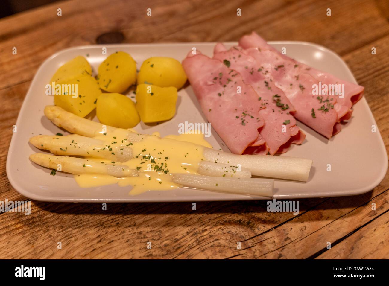 Klassisches Spargelgericht mit Schinken, Kartoffeln und Sauce Hollandaise 17.04.25, Selters: Symbolfoto, Illustrationsbild, Symbolbild, Illustrationsfoto, Alltagsszene Ein traditionelles deutsches Frühlingsgericht mit weißem Spargel, serviert mit Sauce Hollandaise, gekochten Kartoffeln und geräuchertem Kochschinken. DAS Gericht ist angerichtet auf einem hellen rechteckigen Teller auf einem rustikalen Holztisch. Der weiße Spargel ist leicht mit Petersilie garniert, die Kartoffeln sind geschält und in Stücke geschnitten. Eine typische Mahlzeit zur Spargelsaison in Deutschland. . Selters Hessen G. Foto Stock