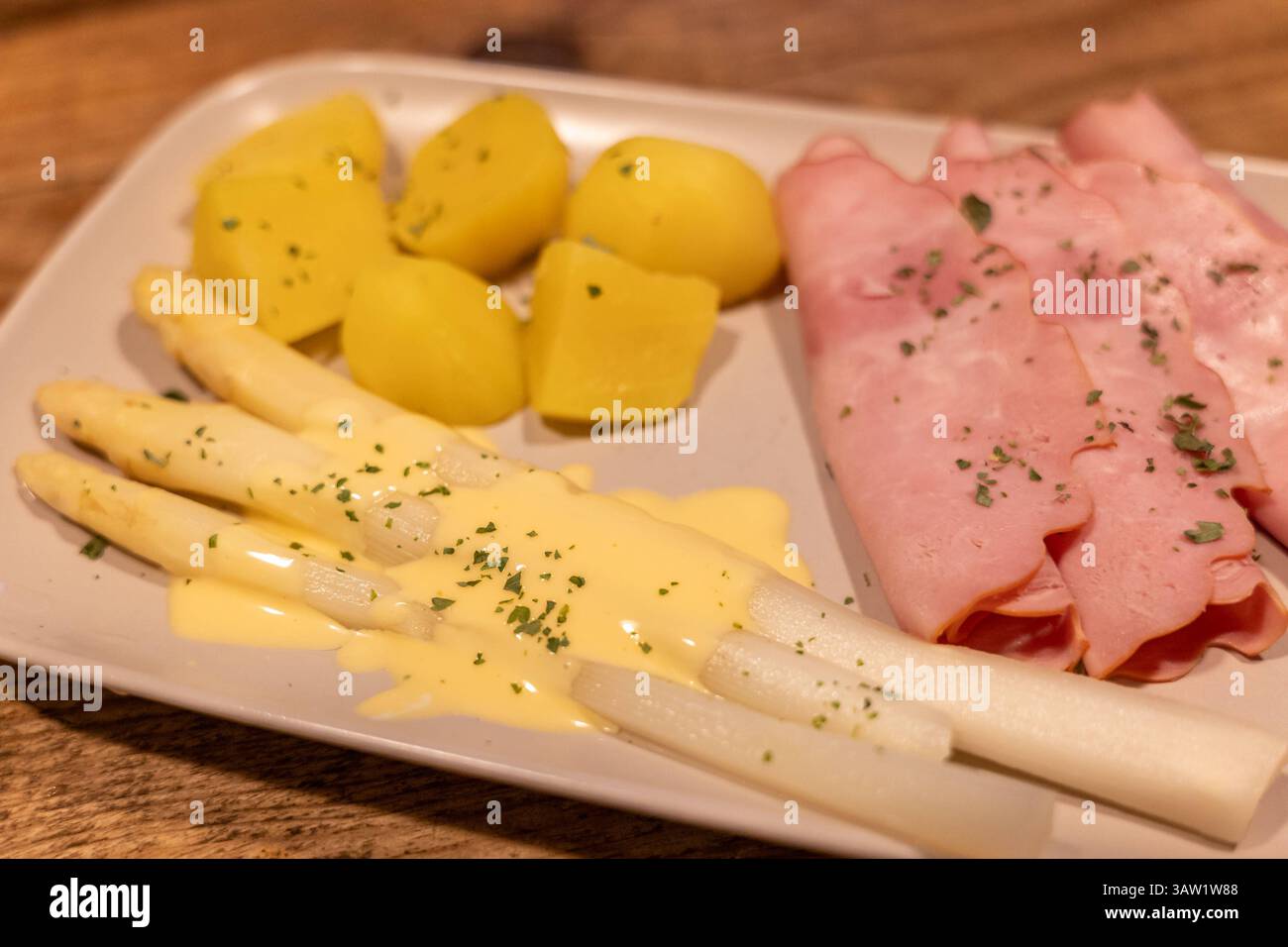 Klassisches Spargelgericht mit Schinken, Kartoffeln und Sauce Hollandaise 17.04.25, Selters: Symbolfoto, Illustrationsbild, Symbolbild, Illustrationsfoto, Alltagsszene Ein traditionelles deutsches Frühlingsgericht mit weißem Spargel, serviert mit Sauce Hollandaise, gekochten Kartoffeln und geräuchertem Kochschinken. DAS Gericht ist angerichtet auf einem hellen rechteckigen Teller auf einem rustikalen Holztisch. Der weiße Spargel ist leicht mit Petersilie garniert, die Kartoffeln sind geschält und in Stücke geschnitten. Eine typische Mahlzeit zur Spargelsaison in Deutschland. . Selters Hessen G. Foto Stock