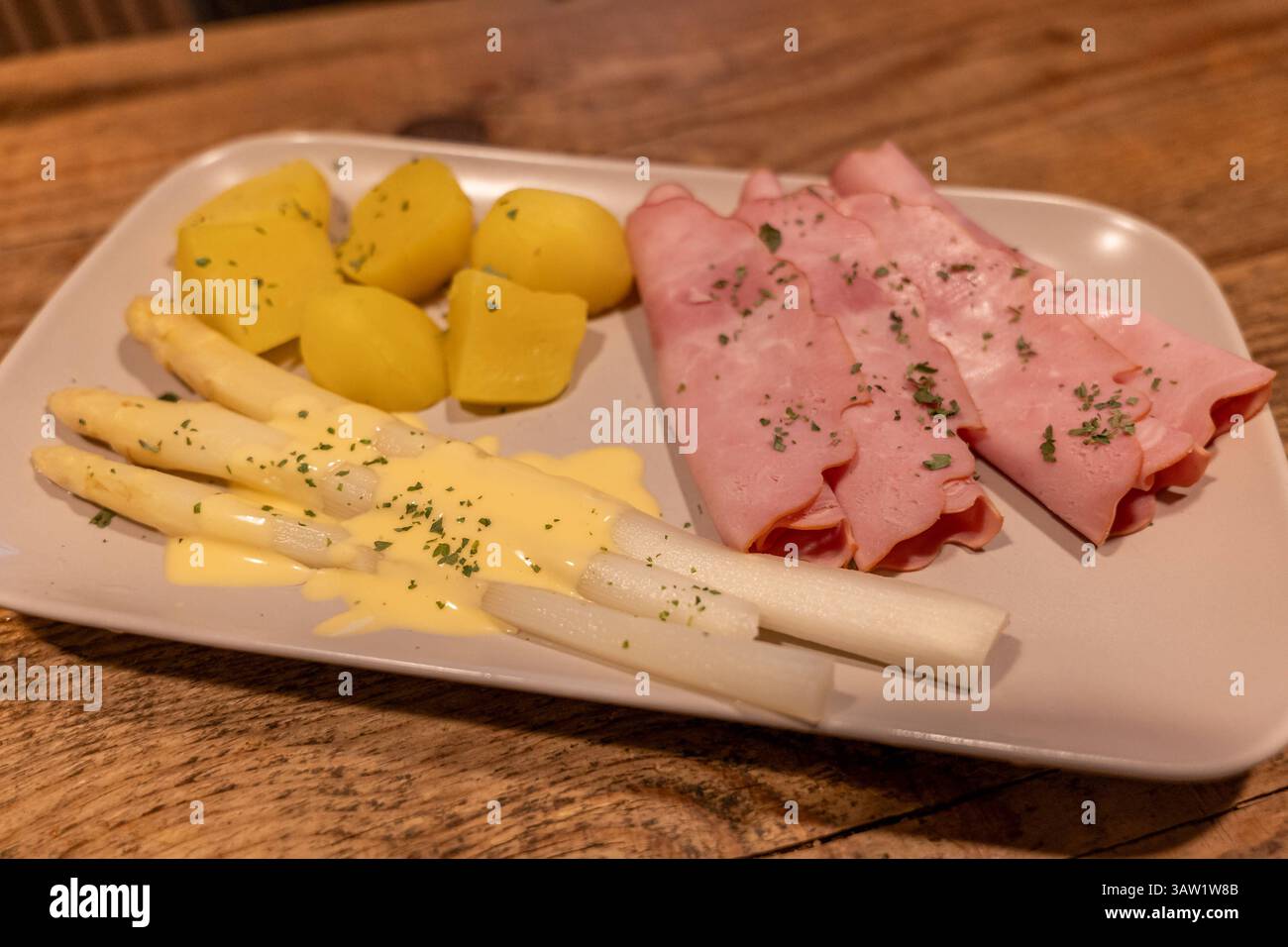 Klassisches Spargelgericht mit Schinken, Kartoffeln und Sauce Hollandaise 17.04.25, Selters: Symbolfoto, Illustrationsbild, Symbolbild, Illustrationsfoto, Alltagsszene Ein traditionelles deutsches Frühlingsgericht mit weißem Spargel, serviert mit Sauce Hollandaise, gekochten Kartoffeln und geräuchertem Kochschinken. DAS Gericht ist angerichtet auf einem hellen rechteckigen Teller auf einem rustikalen Holztisch. Der weiße Spargel ist leicht mit Petersilie garniert, die Kartoffeln sind geschält und in Stücke geschnitten. Eine typische Mahlzeit zur Spargelsaison in Deutschland. . Selters Hessen G. Foto Stock