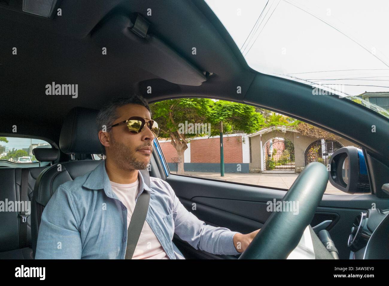 Un uomo che indossa gli occhiali da sole sta guidando un'auto. Indossa una camicia blu e una bianca Foto Stock Un uomo che indossa gli occhiali da sole sta guidando un'auto. Indossa una camicia blu e una bianca Foto Stock