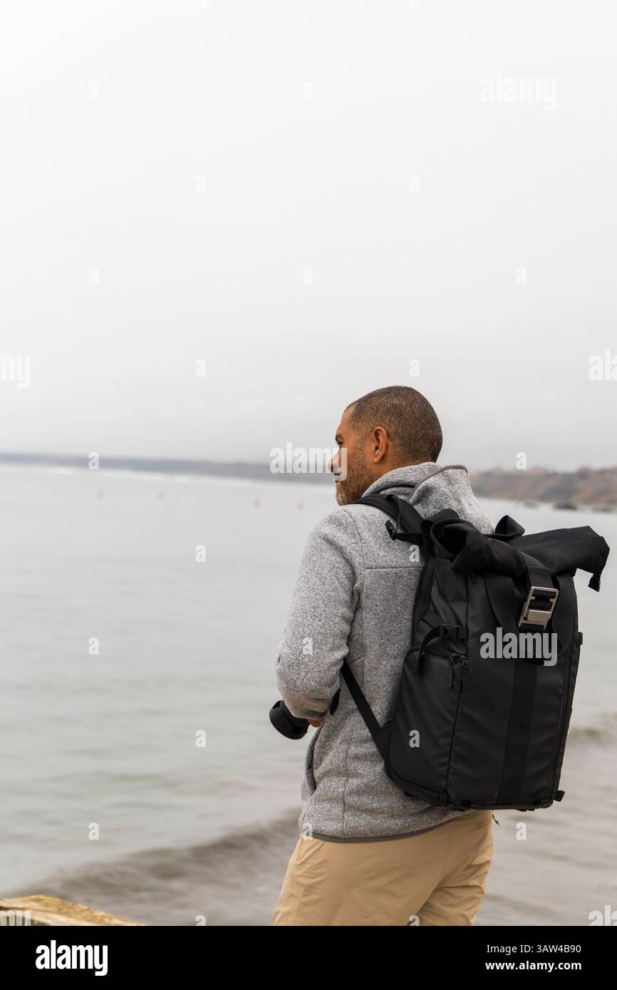 Un uomo è in piedi su una spiaggia con uno zaino sulla schiena. Indossa una giacca grigia e pantaloni kaki. Il cielo è nuvoloso e l'acqua è calma. La ma Foto Stock Un uomo è in piedi su una spiaggia con uno zaino sulla schiena. Indossa una giacca grigia e pantaloni kaki. Il cielo è nuvoloso e l'acqua è calma. La ma Foto Stock