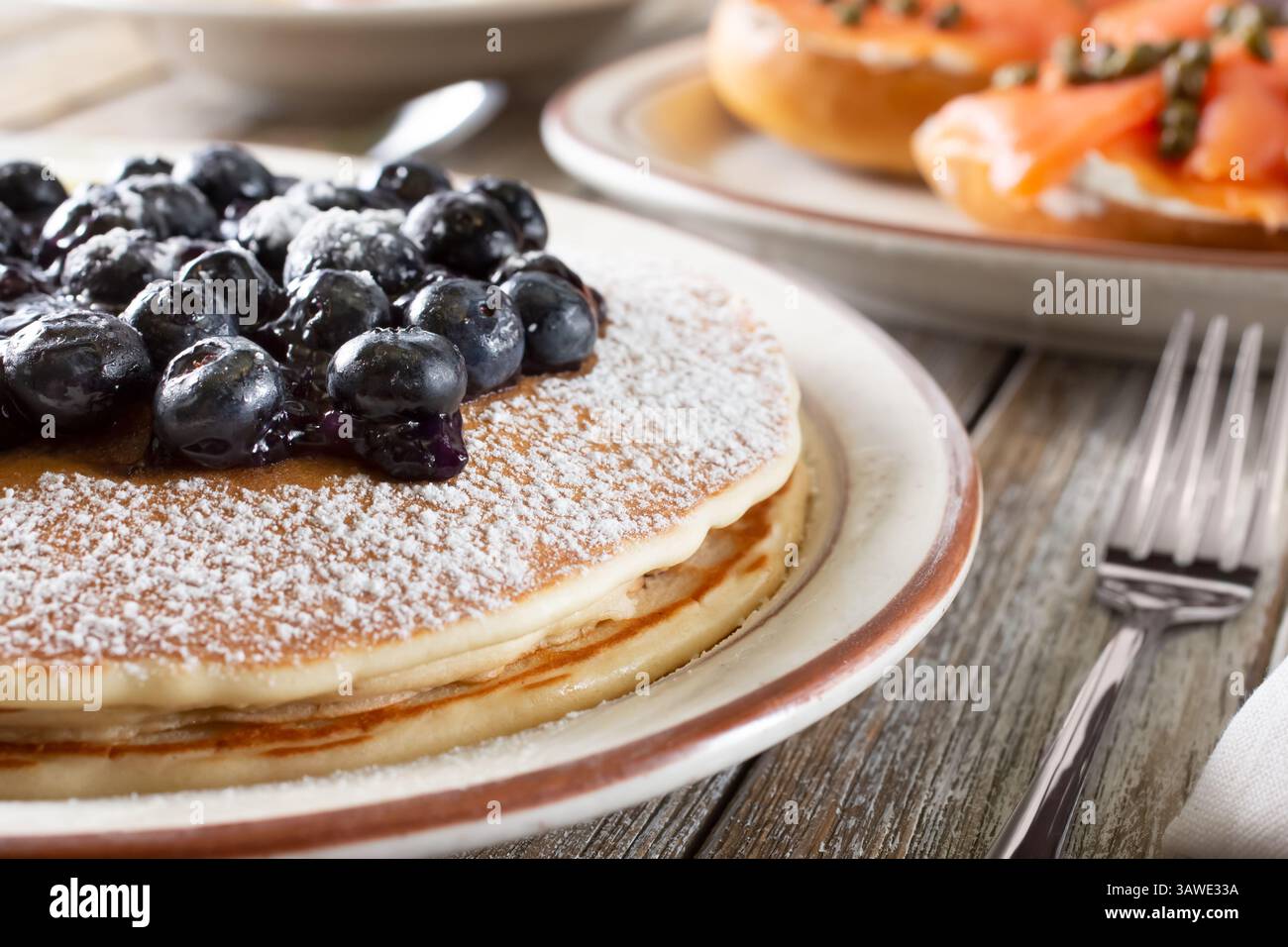 Una vista di un piatto di pancake ricoperti di mirtilli, con un piatto di lox di bagel sullo sfondo. Foto Stock