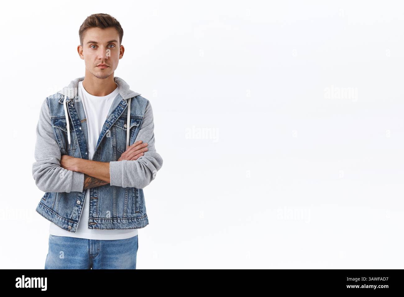 Ritratto del giovane biondo caucasico in giacca denim in piedi su sfondo bianco con espressione normale, guardando fotocamera croce braccia petto sicuro Foto Stock