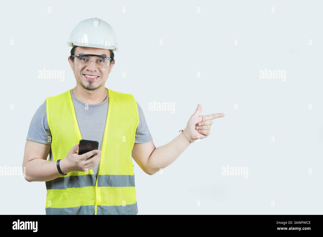 Bell'architetto che tiene il telefono che punta allo spazio vuoto isolato. Giovane ingegnere sorridente che tiene in mano lo smartphone e indica uno spazio vuoto isolato Foto Stock