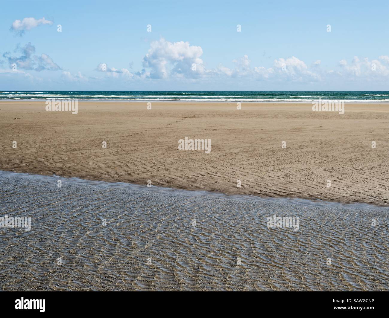 Le onde si infrangono dolcemente contro la riva di Inchydoney Beach, dove la sabbia soffice si estende sotto un cielo azzurro. La luce del sole migliora l'atmosfera serena per Foto Stock