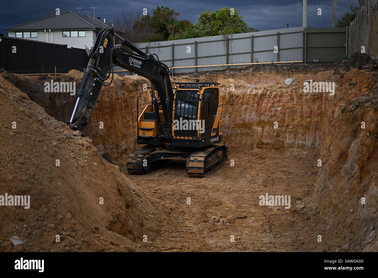 Escavatore cingolato giallo e nero Hyundai HX145LCR seduto in un buco su un cantiere suburbano, circondato da terra rossastra Foto Stock