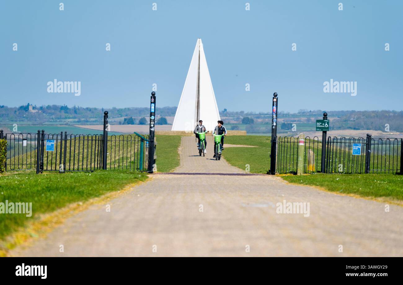 Ragazzi in bicicletta lontano dalla piramide leggera di Milton Keynes. Questa opera d'arte pubblica si trova in cima al tumulo o belvedere, il punto più alto del parco, dove funge da faro per chilometri intorno. È stato acceso per la prima volta per il Giubileo di Diamante della Regina ElizabethÕs nel 2012 ed è ancora utilizzato per commemorare eventi speciali locali e nazionali. Fondata negli anni '1960, Milton Keynes nel Buckinghamshire era una delle "nuove città" del Regno Unito di quell'epoca. Spesso deriso per la sua architettura moderna, la miriade di rotatorie e le famigerate mucche di cemento, è in realtà un posto piuttosto bello da vivere, pieno di molti parchi e passeggiate Foto Stock