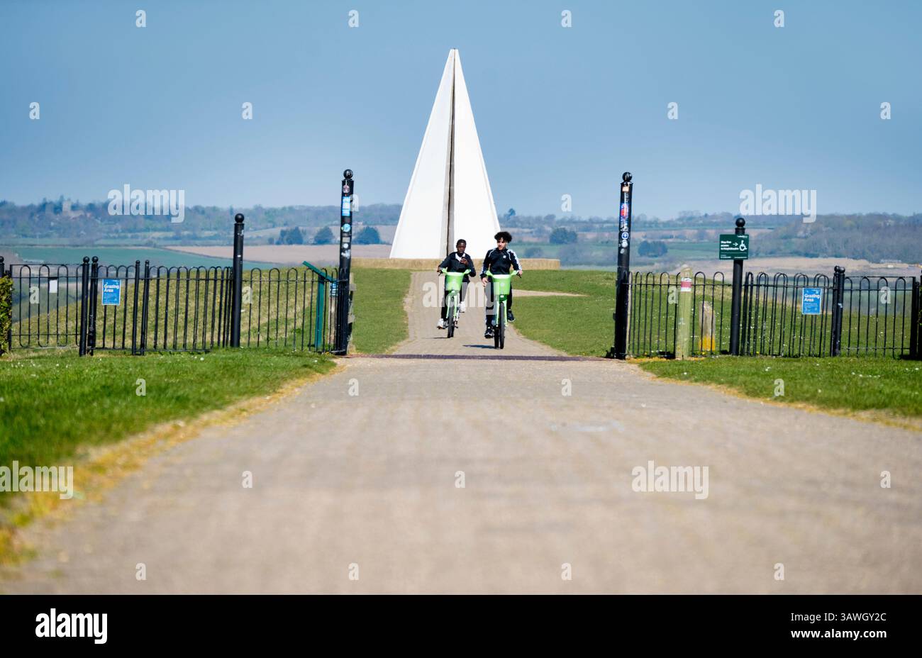 Ragazzi in bicicletta lontano dalla piramide leggera di Milton Keynes. Questa opera d'arte pubblica si trova in cima al tumulo o belvedere, il punto più alto del parco, dove funge da faro per chilometri intorno. È stato acceso per la prima volta per il Giubileo di Diamante della Regina ElizabethÕs nel 2012 ed è ancora utilizzato per commemorare eventi speciali locali e nazionali. Fondata negli anni '1960, Milton Keynes nel Buckinghamshire era una delle "nuove città" del Regno Unito di quell'epoca. Spesso deriso per la sua architettura moderna, la miriade di rotatorie e le famigerate mucche di cemento, è in realtà un posto piuttosto bello da vivere, pieno di molti parchi e passeggiate Foto Stock