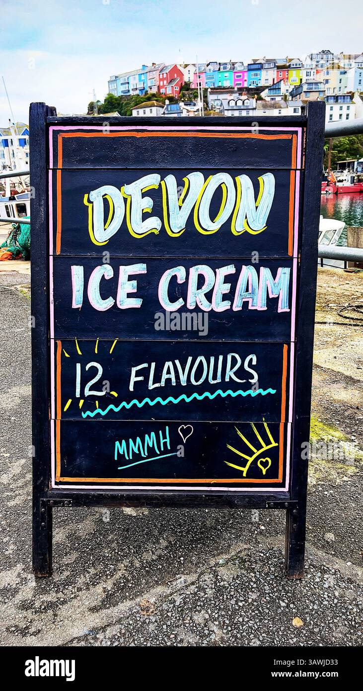 Menu scritto a mano sulla lavagna che mostra una varietà di gusti di gelato, sullo sfondo della costa con barche e porto. Brixham, Devon, Regno Unito Foto Stock