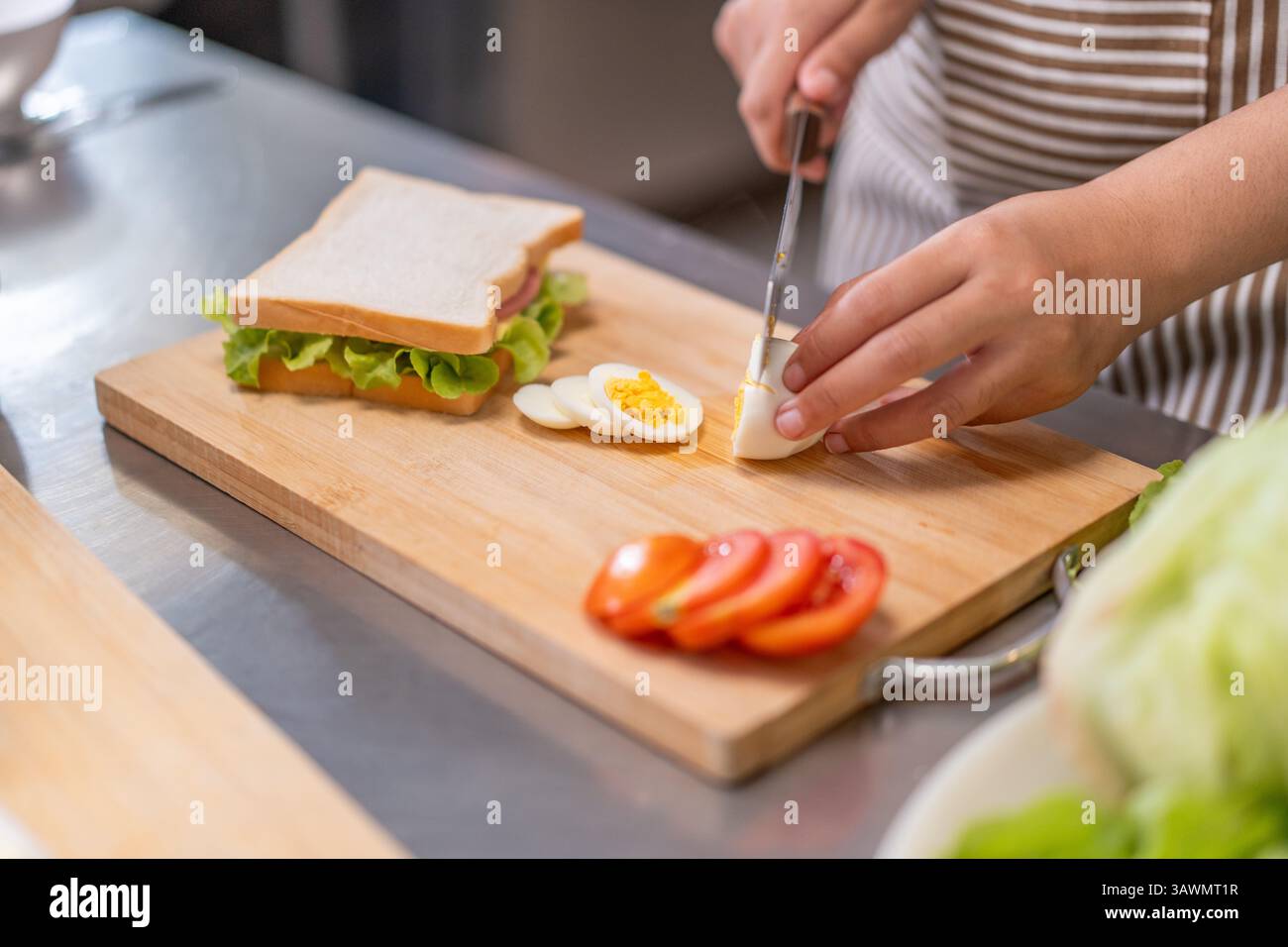Una giovane chef sorridente prepara un panino fresco in una cucina commerciale, circondato da ingredienti, mostrando passione e creatività in cucina. Foto Stock