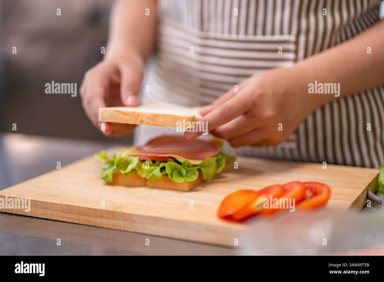 Una giovane chef sorridente prepara un panino fresco in una cucina commerciale, circondato da ingredienti, mostrando passione e creatività in cucina. Foto Stock