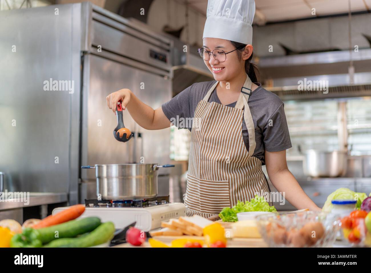La giovane chef donna in grembiule a righe e cappello aggiunge un uovo a una pentola usando un mestolo in una cucina commerciale. Circondata da verdure fresche e pane, lei Foto Stock
