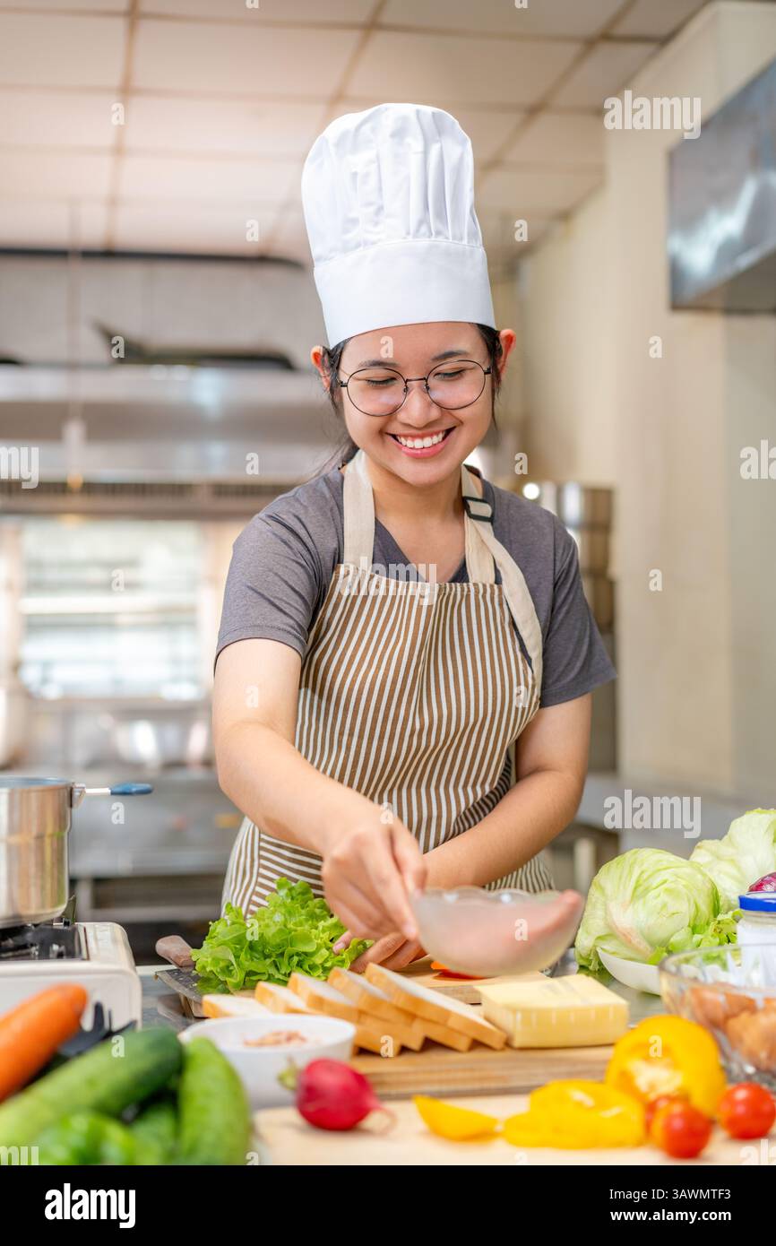 Una giovane chef donna che indossa un grembiule a righe e un cappello da chef che prepara verdure fresche e altri ingredienti in una cucina commerciale. Lei è concentrata whil Foto Stock