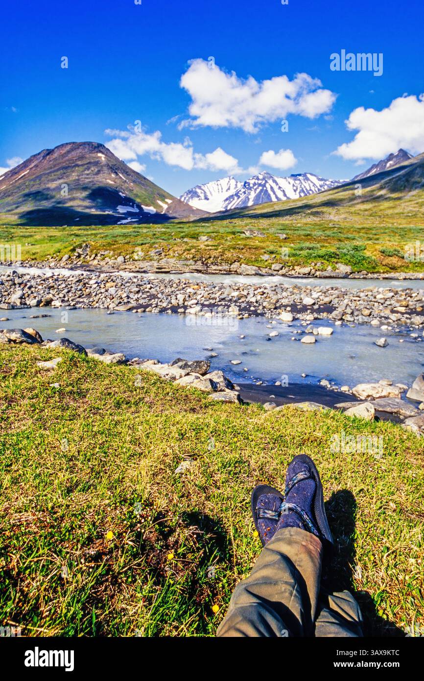 Escursionista che riposa accanto a un fiume in un paesaggio montano panoramico in una giornata estiva di sole Foto Stock