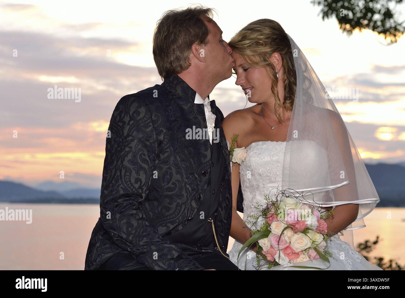 Lo sposo bacia la sposa sulla fronte al tramonto sul lago Foto Stock